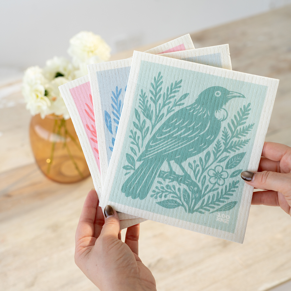 A person holds four Swedish Dishcloths - NZ Woodcut Birds featuring eco-friendly illustrated bird and plant designs in pastel colors. In the background, a vase with white flowers rests on a light wooden table.