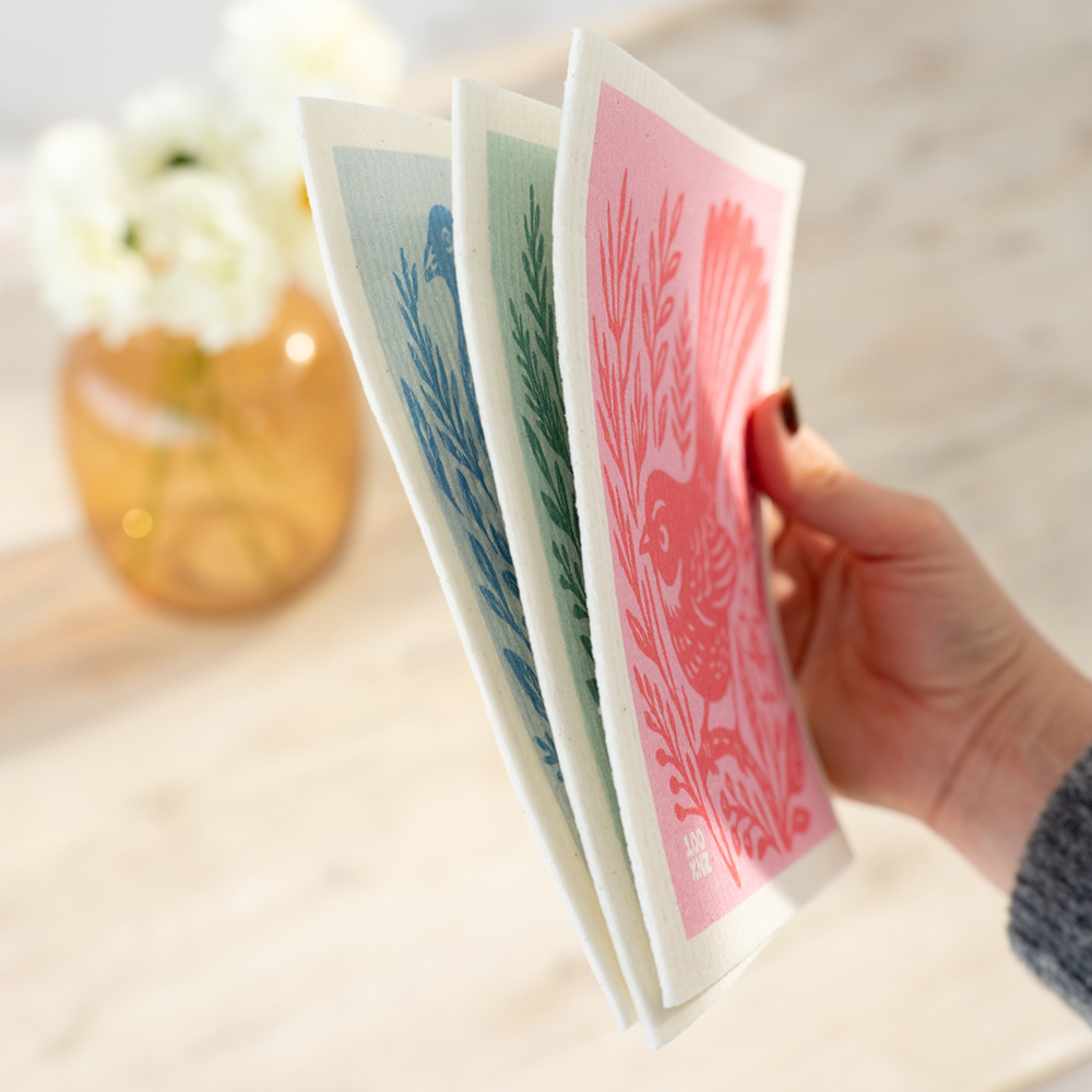 A hand holds three rectangular Swedish Dishcloths - NZ Woodcut Birds, featuring charming bird and plant designs in red, green, and blue. In the background, a vase with white flowers rests on a light wooden surface.