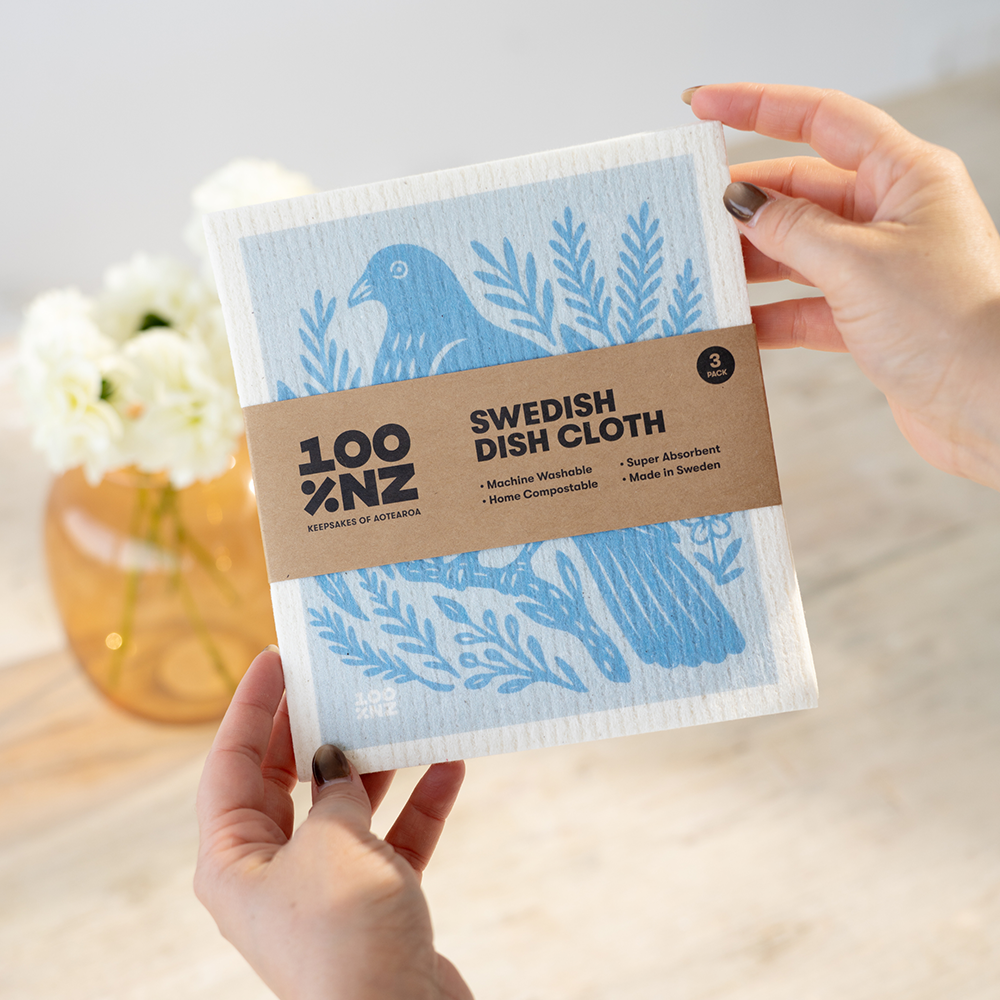 A person holds a pack of Swedish Dishcloths - NZ Woodcut Birds, featuring blue eco-friendly cloths with bird and plant designs. The brown label highlights features such as washable, home compostable, absorbent, and made in Sweden. A flower vase appears in the background.
