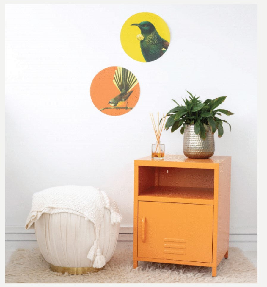 An orange side table with a plant and reed diffuser sits next to a white pouf with blanket. Two Bright Huia Art Spot prints hang above on the wall, while a rug covers the floor.