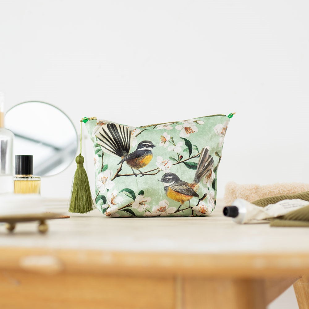 The Chatty Fantails Velvet Cosmetic Bag, designed in New Zealand with floral prints and illustrated birds, sits on a wooden surface among beauty essentials—making it a perfect gift.
