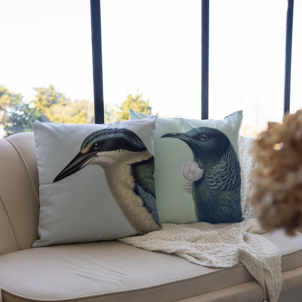 Two Hushed Green Tui Cushion Covers, featuring detailed bird illustrations inspired by New Zealand native birds, rest on a light beige sofa near a large window. A knit throw is draped across the sofa, while treetops can be seen outside. Soft daylight gently highlights each cushion cover’s unique New Zealand design.