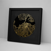 The Auckland Mapscape Foil Print, a black-framed picture with gold map lines on a black background, brings contemporary flair to white decor, displayed upright against a light wall and resting on a white surface.