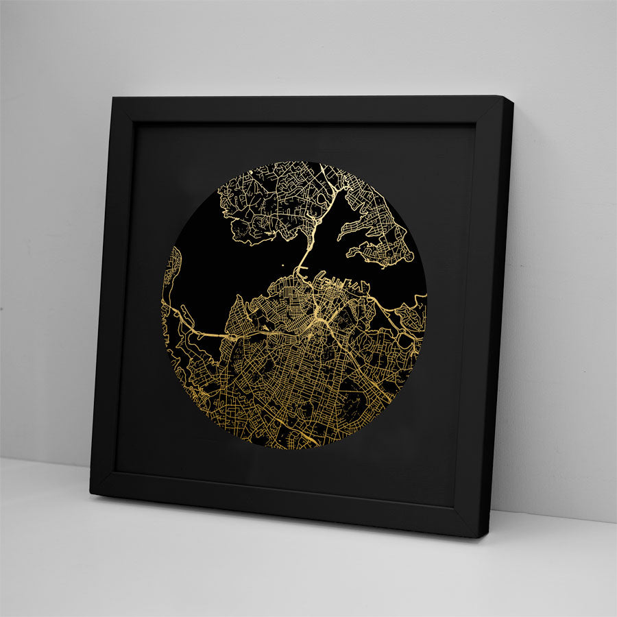 The Auckland Mapscape Foil Print, a black-framed picture with gold map lines on a black background, brings contemporary flair to white decor, displayed upright against a light wall and resting on a white surface.