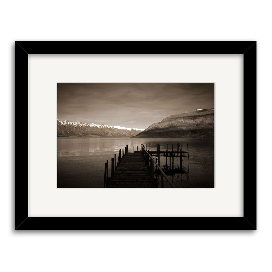 Lake Wakatipu Wharf - RP066: A black and white framed photo of a wooden pier stretching into a serene New Zealand lake, mountains beyond under cloudy skies. Available as photographic prints on archival quality matte paper.