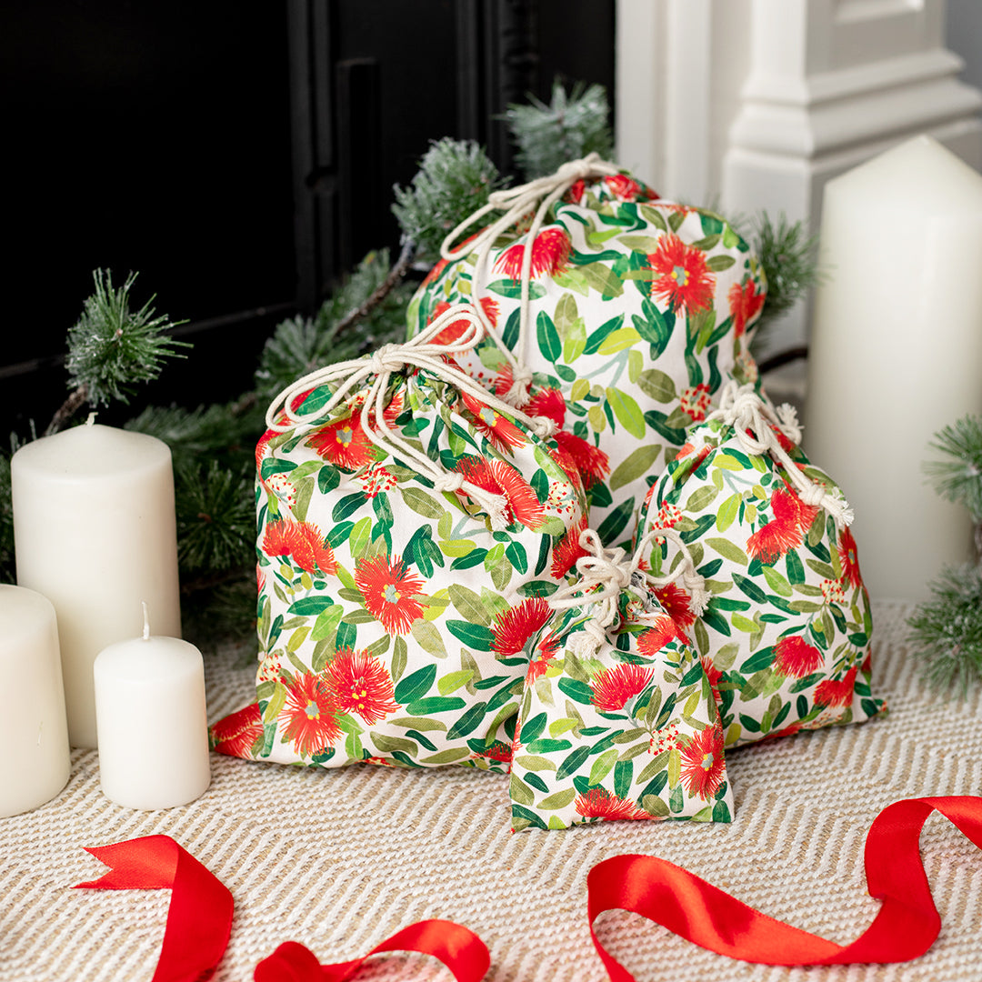 The Pohutukawa Print Drawstring Bag Set features four reusable bags with a red and green floral pattern, displayed on a woven surface with seasonal decor in front of a fireplace.