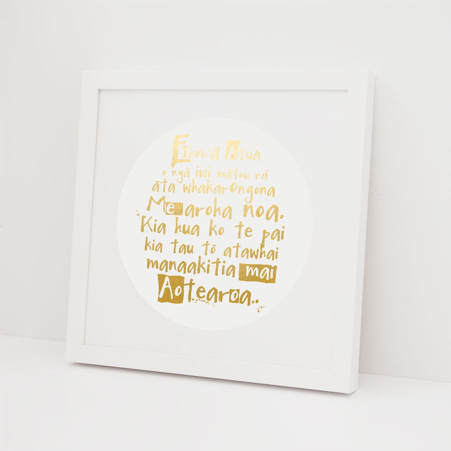 The National Anthem Foil Print features gold te reo Māori text in a circular design on white, with a white frame and slight left lean—a unique Kiwiana art piece available as framed or unframed gold foil print.