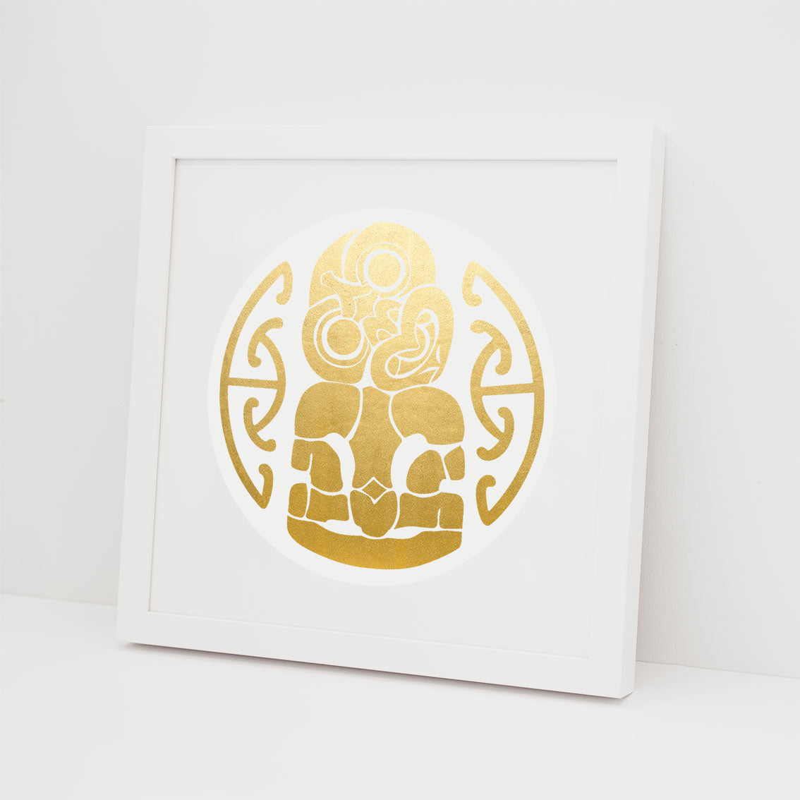 The Tiki Foil Print is a framed gold foil artwork with a symmetrical Maori-inspired design on a white background. This striking piece of Kiwiana art is proudly made in New Zealand, perfect for adding cultural flair to any space.