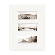 The Auckland Triptych - RP004 is a white-matted set of three archival sepia photographic prints showing New Zealand landscapes: a plant-lined coastline, a sailboat marina, and a rocky shore with foliage.