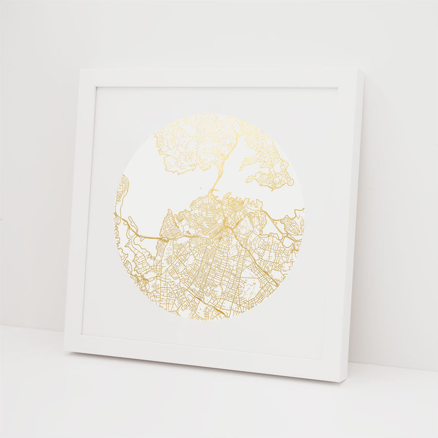 The Auckland Mapscape Foil Print is a modern white-framed picture showcasing a circular gold-foil city map on a white background, leaning against a white wall—ideal for fans of elegant gold foil prints.