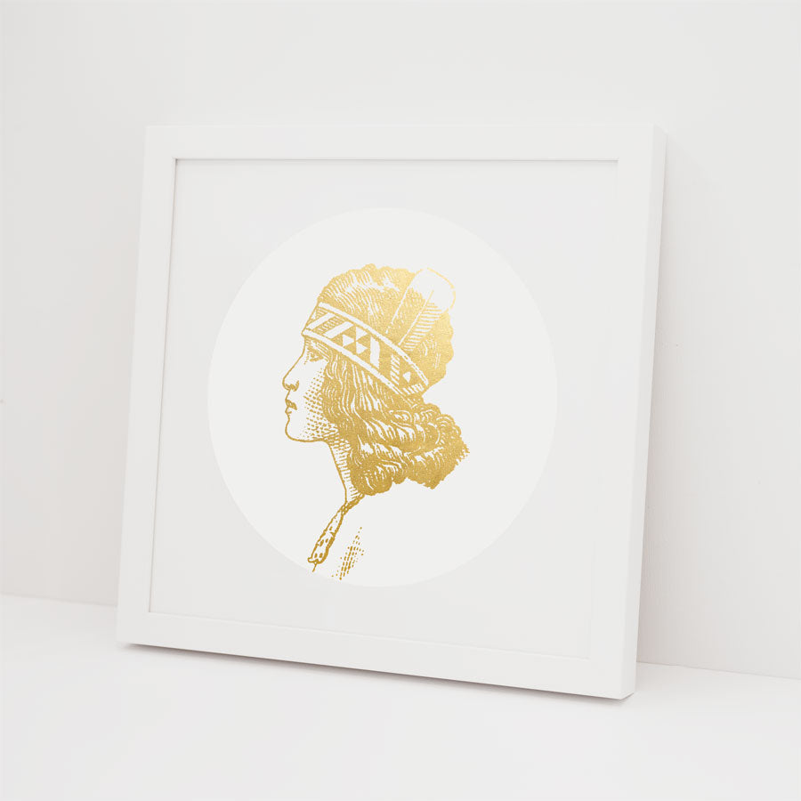 The Wahine Foil Print is a contemporary white framed artwork showcasing a gold foil illustration of a profile in a feathered headdress on a plain background.