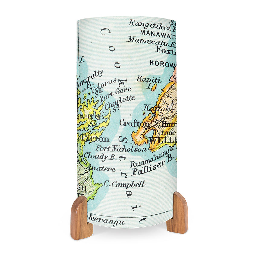 The Vintage Cook Strait Map Table Lamp features a cylindrical shade with vibrant full-colour images of a vintage New Zealand map, displaying place names like Palliser, Port Nicholson, and Manawatu. It has wooden legs and a light blue background.