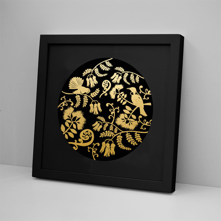 The NZ Design Foil Print features a black frame and circular gold foil prints of birds, flowers, and leaves on a black background, leaning against a light-colored wall.