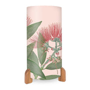 The Vintage Botanical Pohutukawa Table Lamp features a cylindrical shape with a pink background, vibrant red flower and green leaf designs, and simple wooden feet.