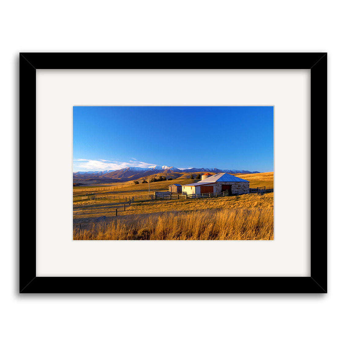 Central Otago - RP074 is a framed photographic print by Reuben Price featuring a small house, outbuilding, golden New Zealand field, snow-capped mountains, clear blue sky, and a wooden fence in the foreground.