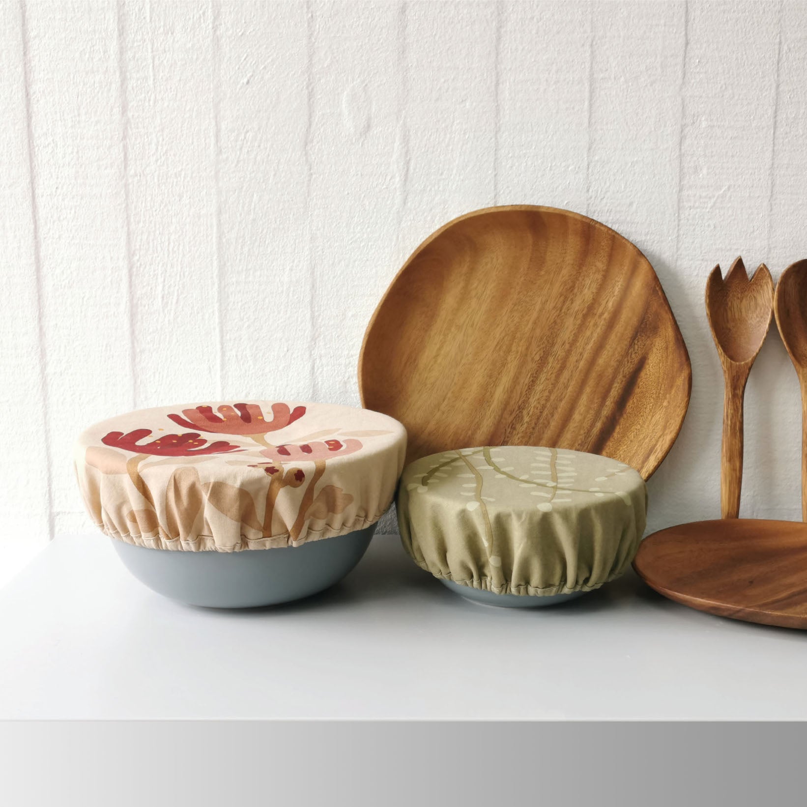 The Raw Bowl Covers (set of 2), featuring food-safe lining and cotton outer, are shown on ceramic bowls beside a serving spoon, spork, and small dish—all displayed on a white surface against a textured white wall.