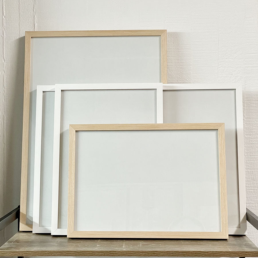 Blank Frames: Set of four blank frames in assorted sizes—two wooden and two white—stand upright and overlap on a wooden surface against a textured white wall, ideal for DIY framing or use with your favorite hanging kit.