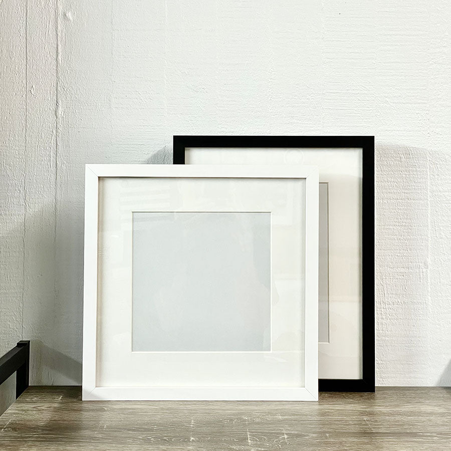 Blank Frames in white and black are displayed on a wooden surface against a white textured wall, with the white frame positioned slightly in front—ideal for your next DIY framing project.