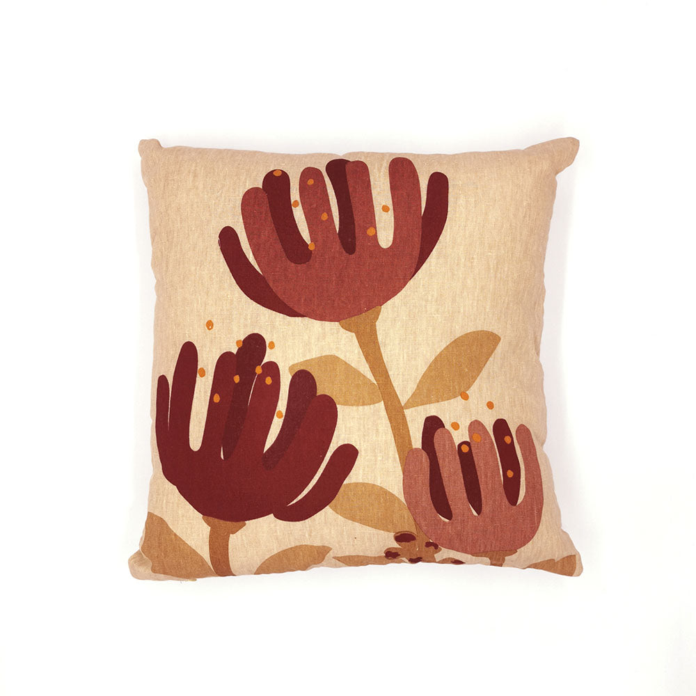 The Raw Pohutukawa Linen Cushion Cover is a square beige throw pillow in natural linen, decorated with three stylized maroon and brown pohutukawa flowers with stems and leaves on the front, set against a plain white background.