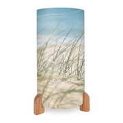 The Beach Grass Table Lamp features a cylindrical shade with a beach grass and sand motif against a blue sky, inspired by New Zealand’s landscapes. It stands on two wooden supports at the base for a stylish, natural touch.