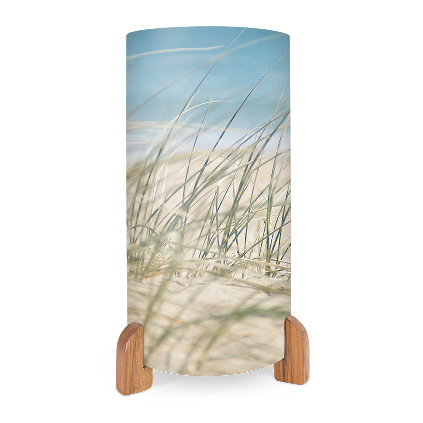The Beach Grass Table Lamp features a cylindrical shade with a beach grass and sand motif against a blue sky, inspired by New Zealand’s landscapes. It stands on two wooden supports at the base for a stylish, natural touch.