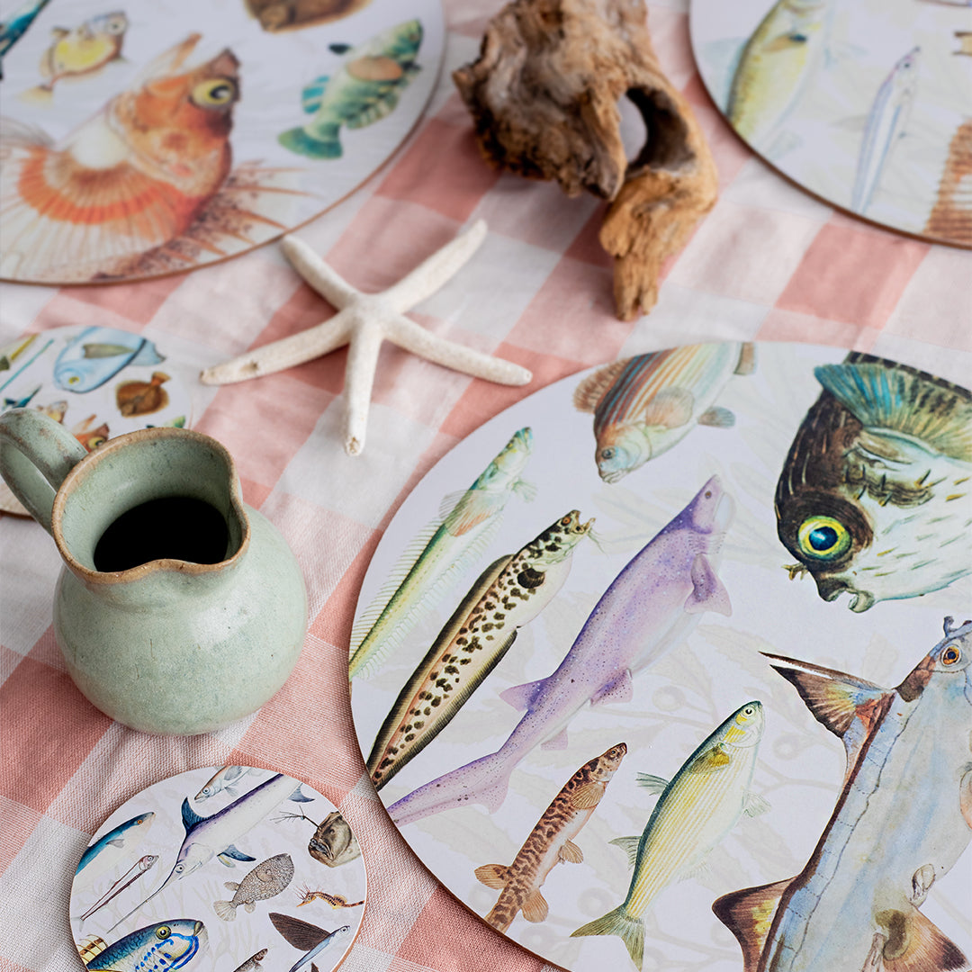 A table is set with the Fishes of NZ Box of 6 Placemats featuring fish illustrations, a matching coaster, a ceramic jug, starfish, and driftwood on a pink and white checkered tablecloth.