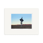 A small art print titled To Falls Dam by New Zealand artist Grahame Sydney, featuring a black road sign, a small post, dry grassy hills, and a clear blue sky.