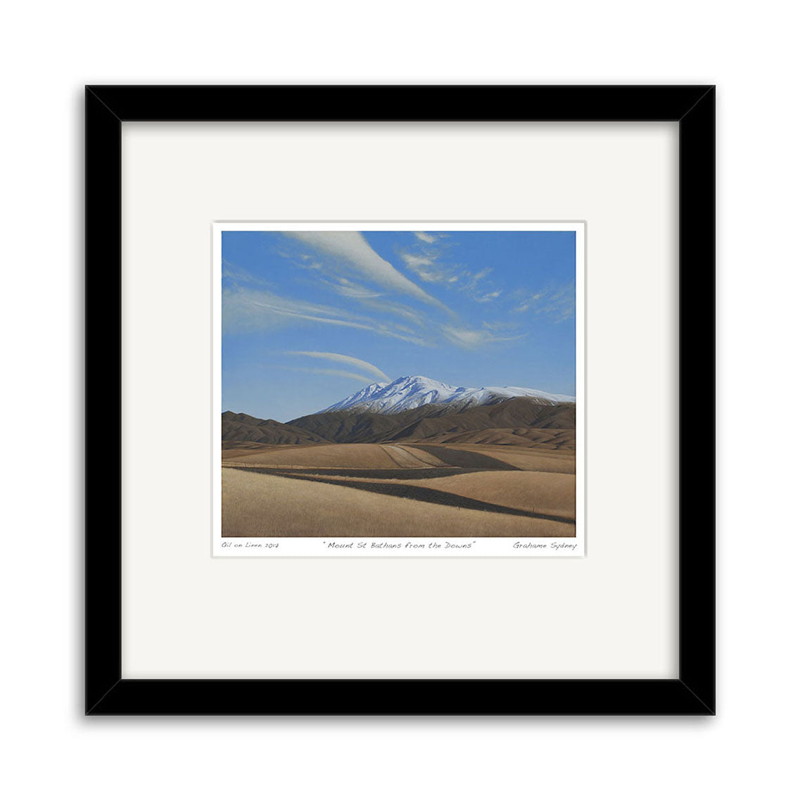 The Grahame Sydney - Mt St Bathans from the Downs Small Art Print shows a snow-capped mountain beneath a blue sky with wispy clouds, rolling brown hills, and desert terrain, all framed in black with a white mat.