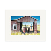A figure stands in the doorway of a weathered red barn with a tractor inside, set among tall grass and open fields beneath a clear sky in Graham Youngs The Red Barn small art print.