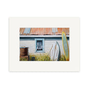 Graham Youngs The Back of the Bach Small Art Print features a weathered white house with a rusted roof, two surfboards, a barrel, and tall grass—an ideal addition to any art collection.
