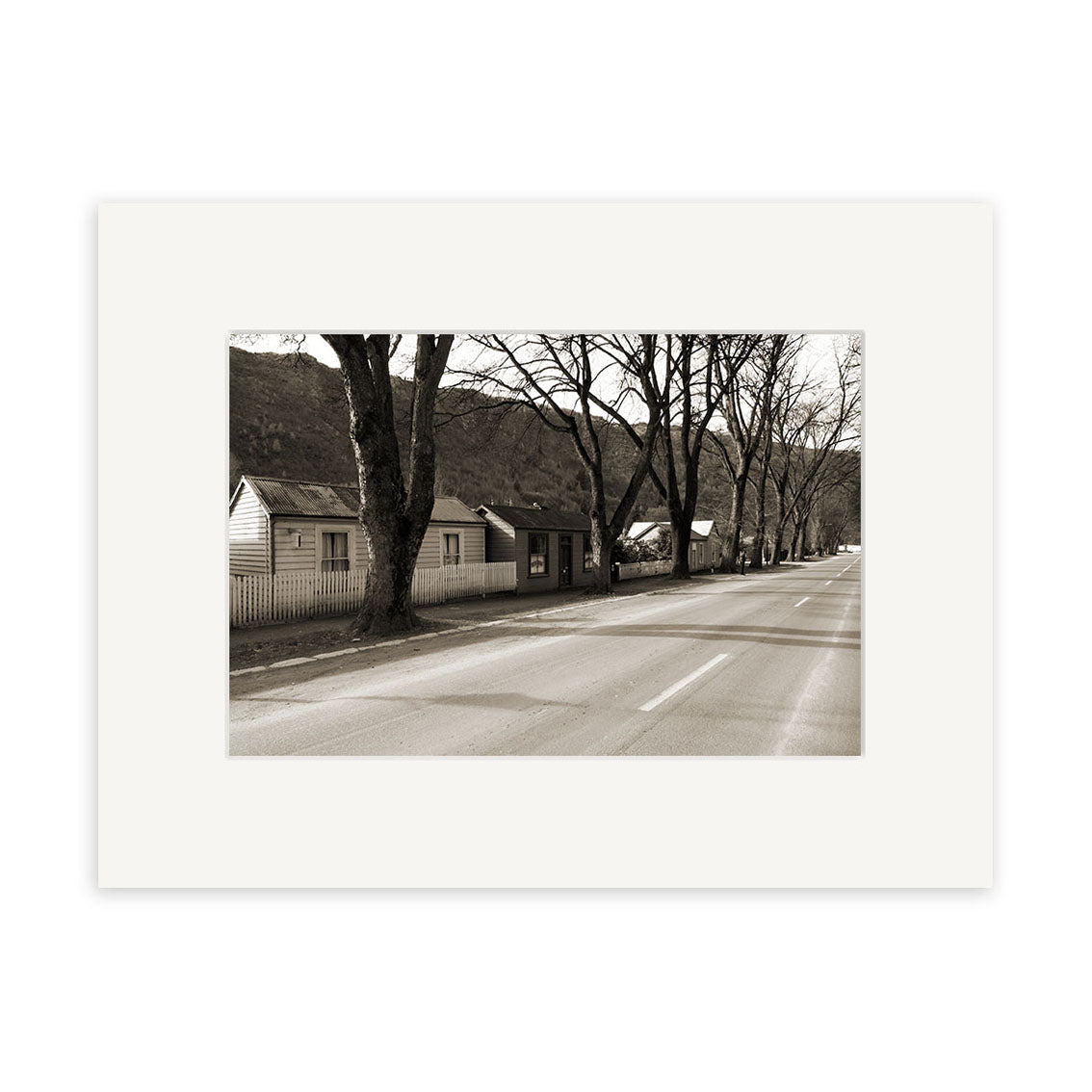 Arrowtown - RP068 features a sepia-toned photographic print of a quiet New Zealand street with leafless trees, small houses, and a distant mountain, showcasing archival quality preservation.