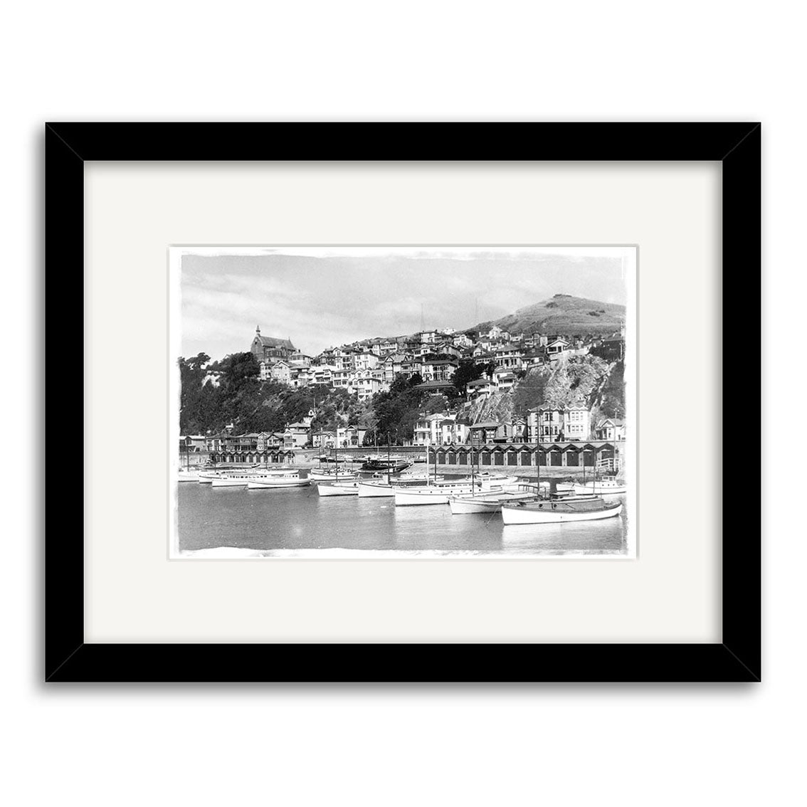 Oriental Bay 1929 - HI001: Black and white historical photo print of a New Zealand coastal town with hillside buildings, boats, and a church. Framed with a white mat and black frame on archival-quality matte paper.