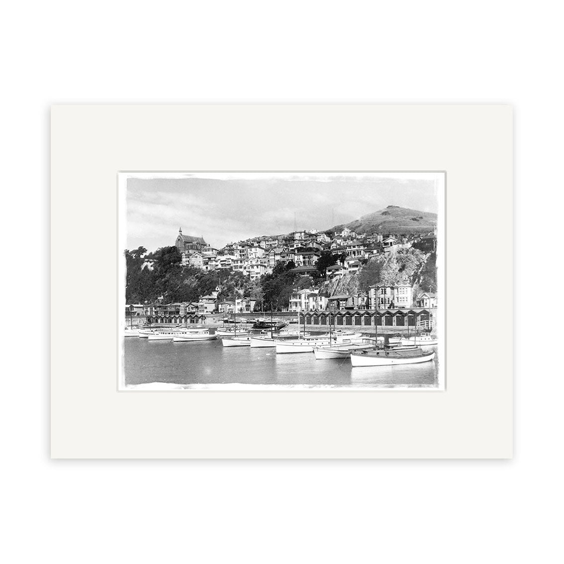 Oriental Bay 1929 - HI001: Black and white photo of a New Zealand coastal town with docked boats, hillside buildings, and a church. Presented as a historical photographic print on archival matte paper with white mat border.