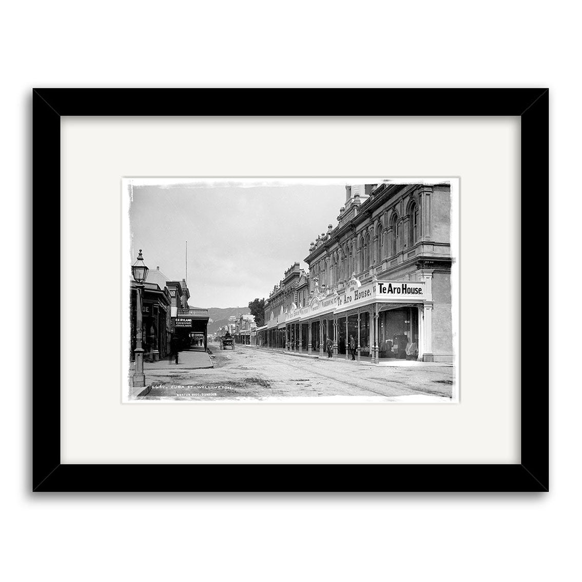 Cuba St 1888 - HI002: Black-and-white historical photographic print of a New Zealand city street with multi-story buildings and Te Aro House sign, framed with white mat and black frame, printed on archival quality matte paper.