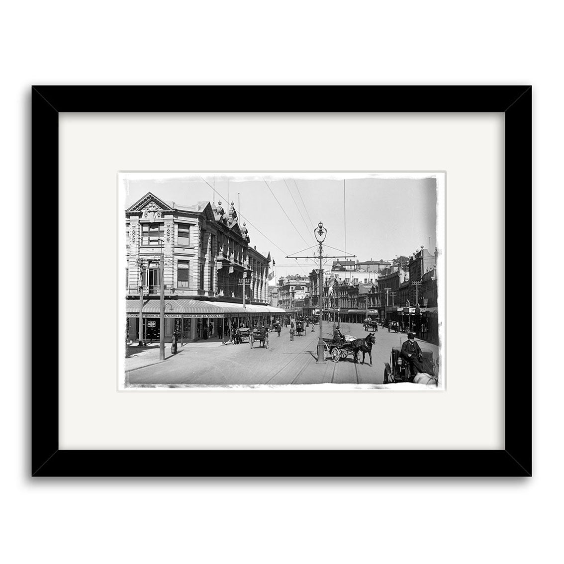 Lambton Quay 1900 - HI003: Archival quality black-and-white print of a bustling New Zealand city street with horse-drawn carriages and historic buildings, framed with a wide white mat and black frame.