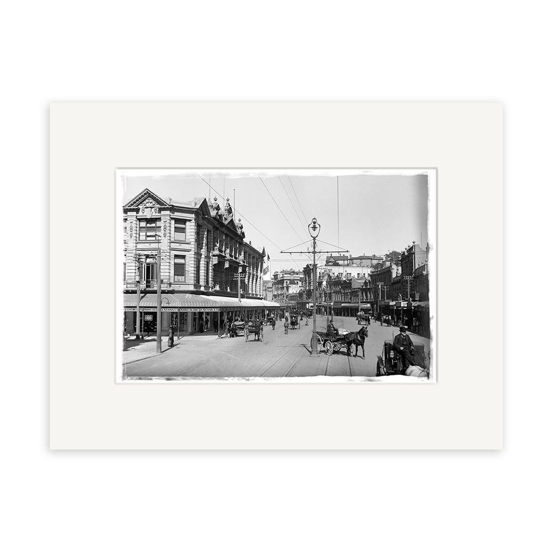 Lambton Quay 1900 - HI003: Archival black and white photo of a New Zealand city street with horse-drawn carriages, pedestrians, multi-story buildings, and an old streetlight.