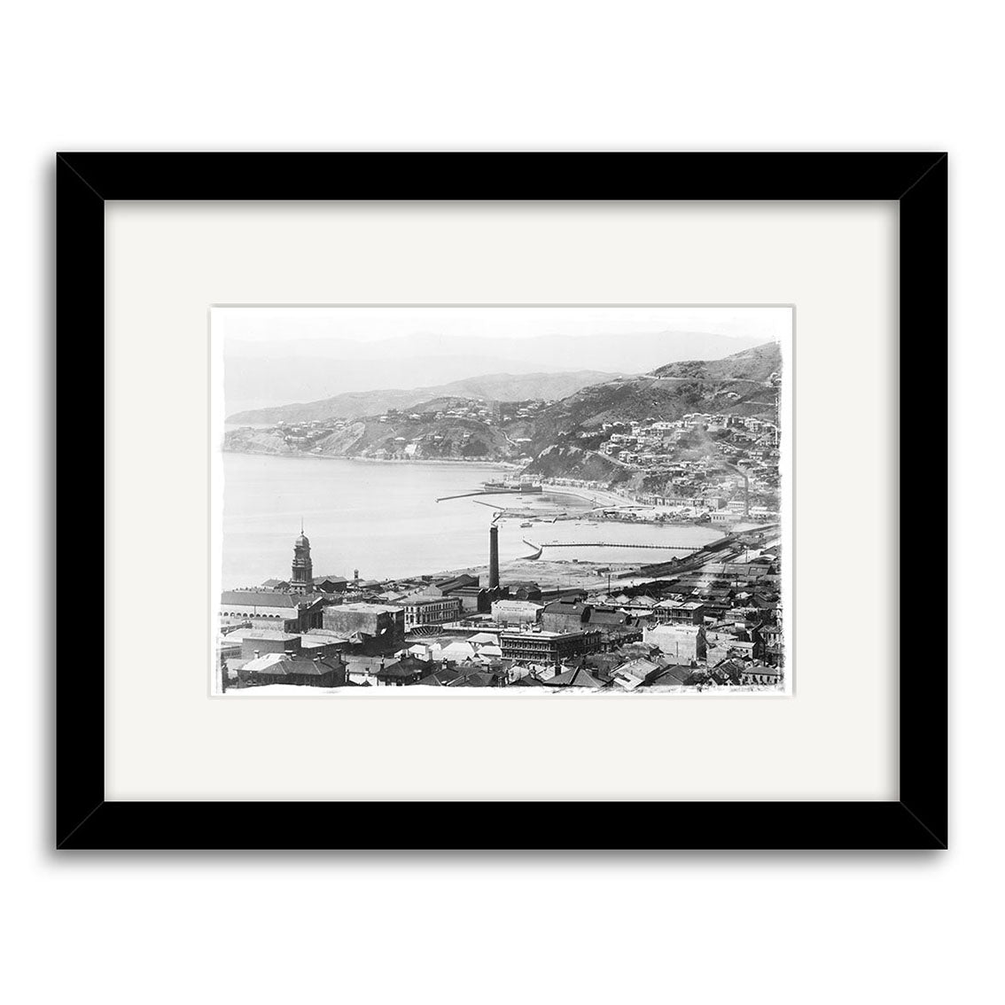 Black-and-white historical photographic print, Wellington City 1905 - HI004, shows a coastal New Zealand city with mountains, buildings, and a tower, beautifully framed with a white mat and black frame for archival quality.