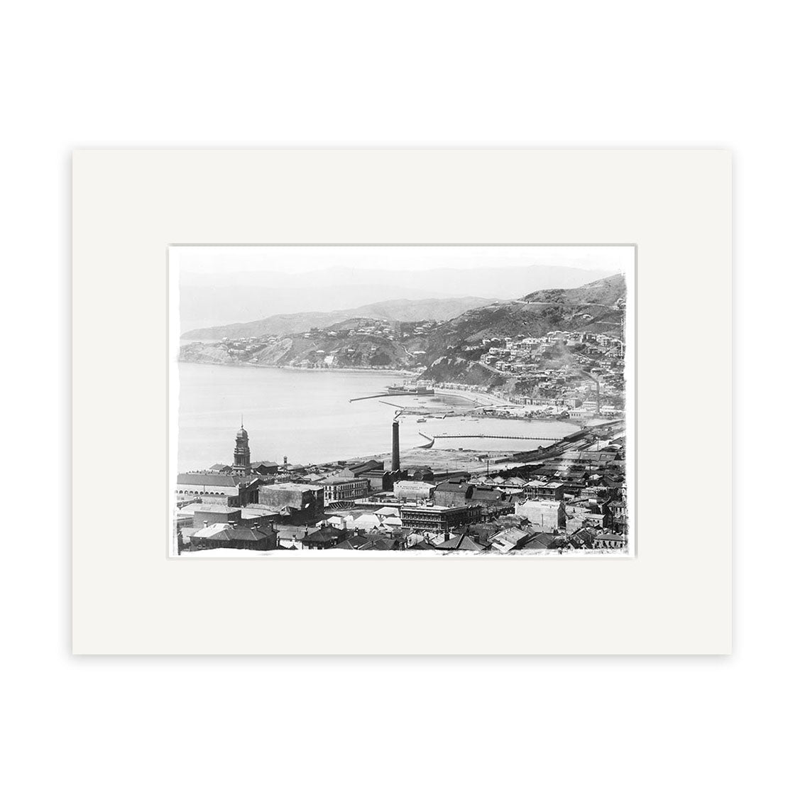 Wellington City 1905 - HI004: Black and white historical photographic print of coastal Wellington with hills, buildings, a tower, bay, docks, and curved shoreline; image is surrounded by a white mat.