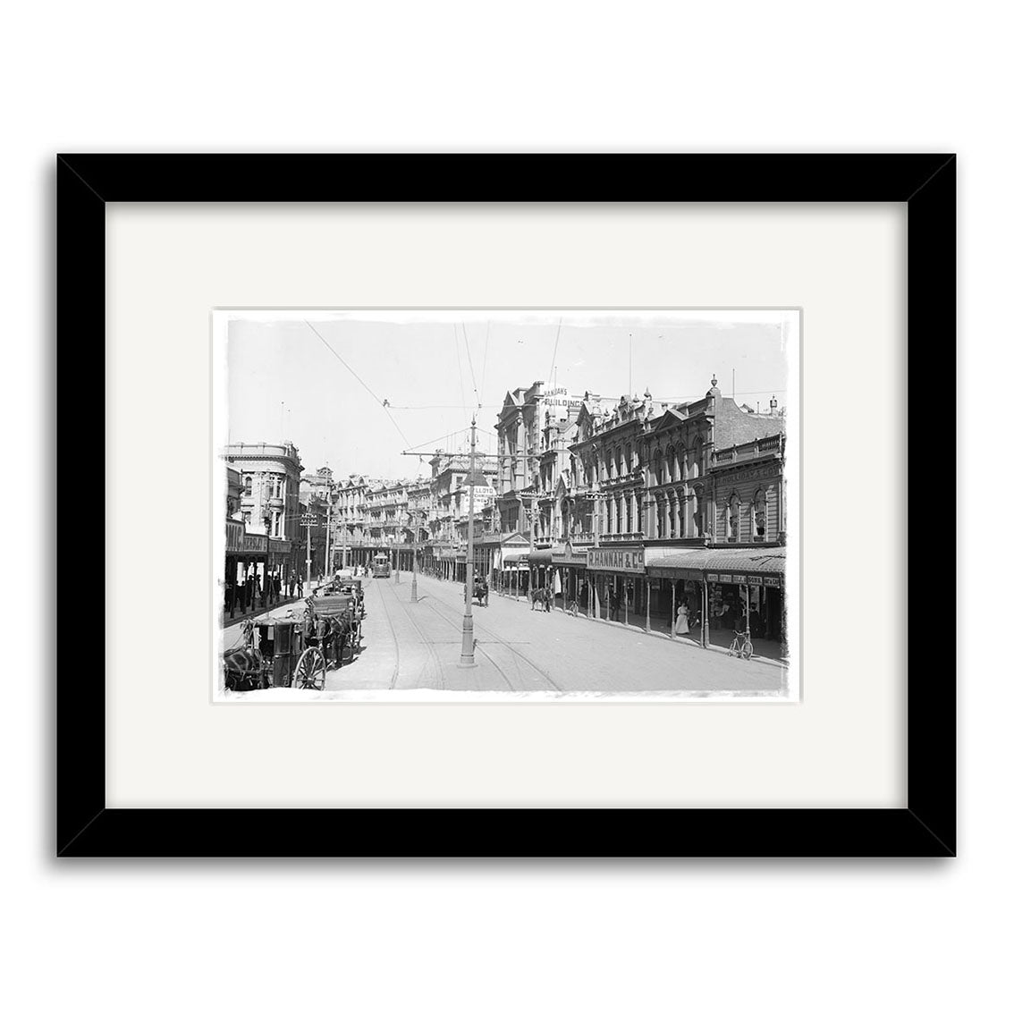 Lambton Quay 1900 - HI005: Archival-quality black & white photo of a New Zealand street with historic buildings, streetcar, and overhead wires, framed in black with a white mat.