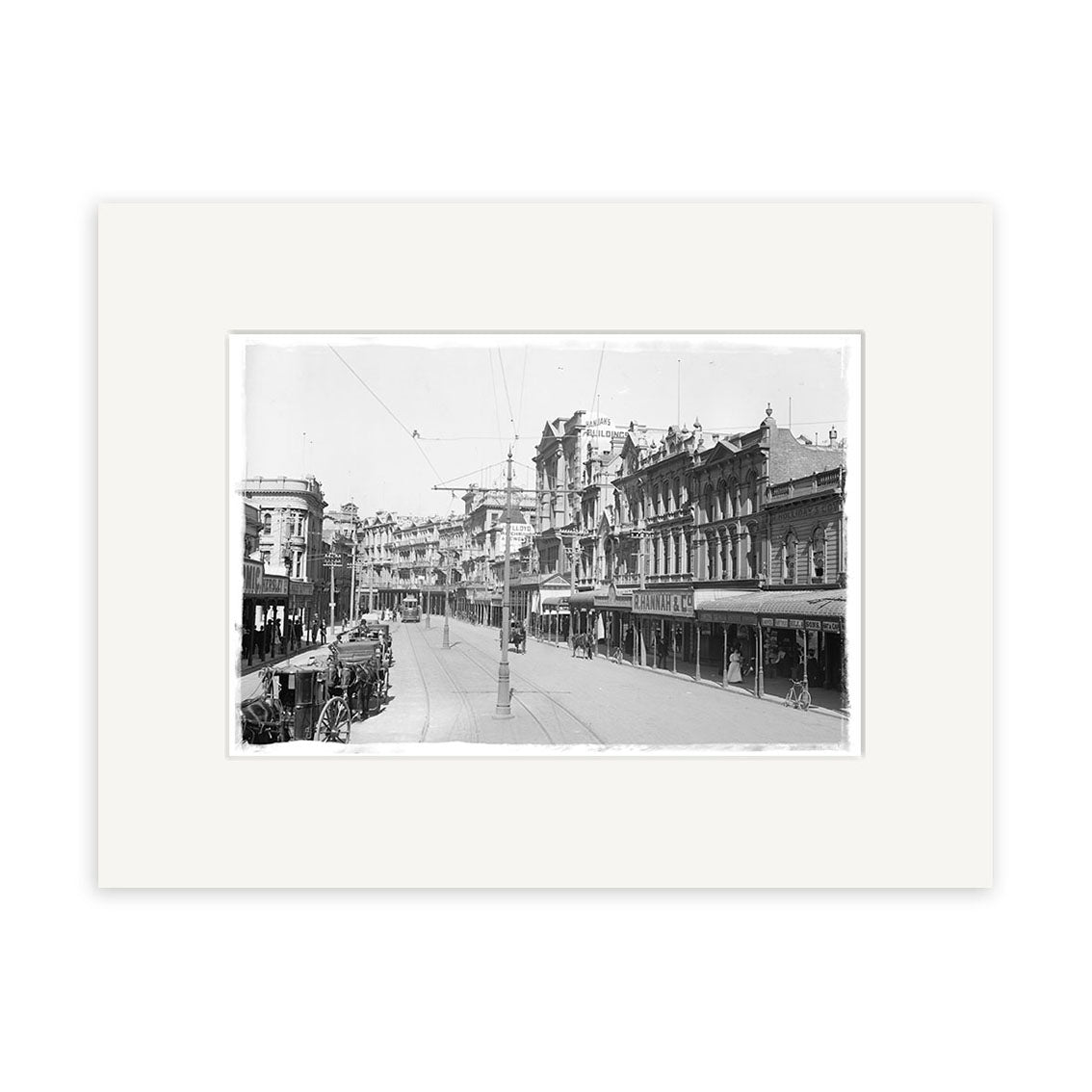 Lambton Quay 1900 - HI005: Black-and-white photo of a New Zealand city street with historic buildings, streetcar tracks, horse-drawn carts, storefronts, and overhead power lines. Matted with a white border.