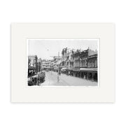 Lambton Quay 1900 - HI005: Black-and-white photo of a New Zealand city street with historic buildings, streetcar tracks, horse-drawn carts, storefronts, and overhead power lines. Matted with a white border.