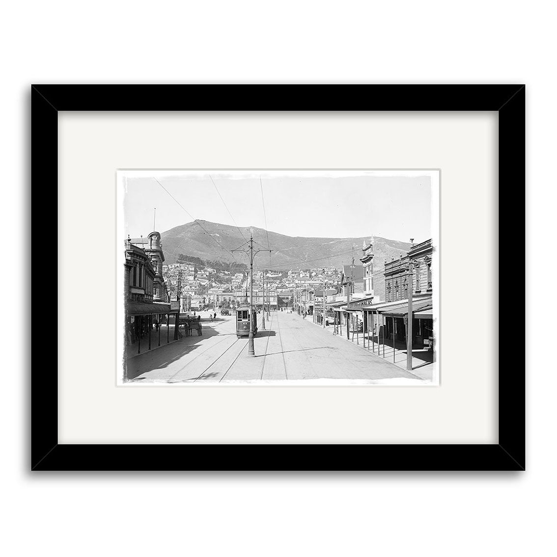 “Courtenay Place 1900 No.1 - HI006” is a black-and-white historical print featuring a wide street with trolley tracks, buildings, and a mountain in New Zealand. Framed with a white mat and black frame on archival matte paper.