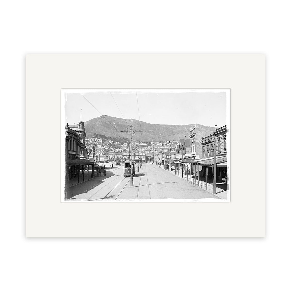 Black-and-white photo titled Courtenay Place 1900 No.1 - HI006 shows a wide New Zealand street with trolley tracks, buildings, a mountain backdrop, few pedestrians, and streetcars. Historical Photographic Print on archival matte paper.