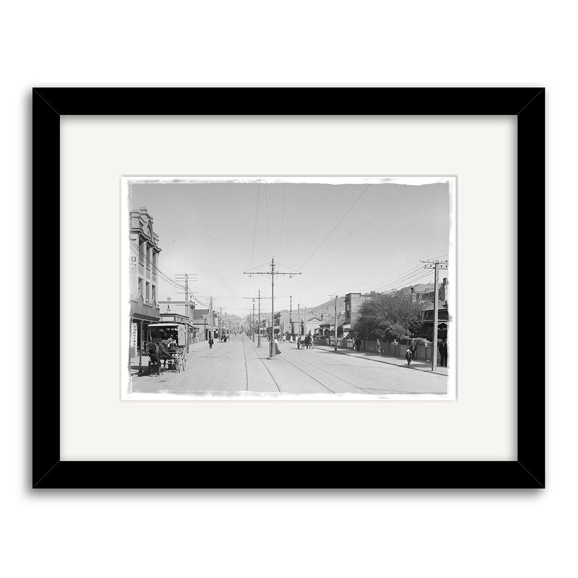 Archival quality Courtenay Place 1900 No.2 - HI007 black and white photo print of a New Zealand street with tram tracks, pedestrians, horse-drawn carts, and shops; framed with a white mat and black border.