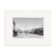 Archival quality black-and-white photographic print: Courtenay Place 1900 No.2 - HI007 shows a New Zealand city street with tram line, utility poles, buildings, and pedestrians, framed by a white mat for preservation.
