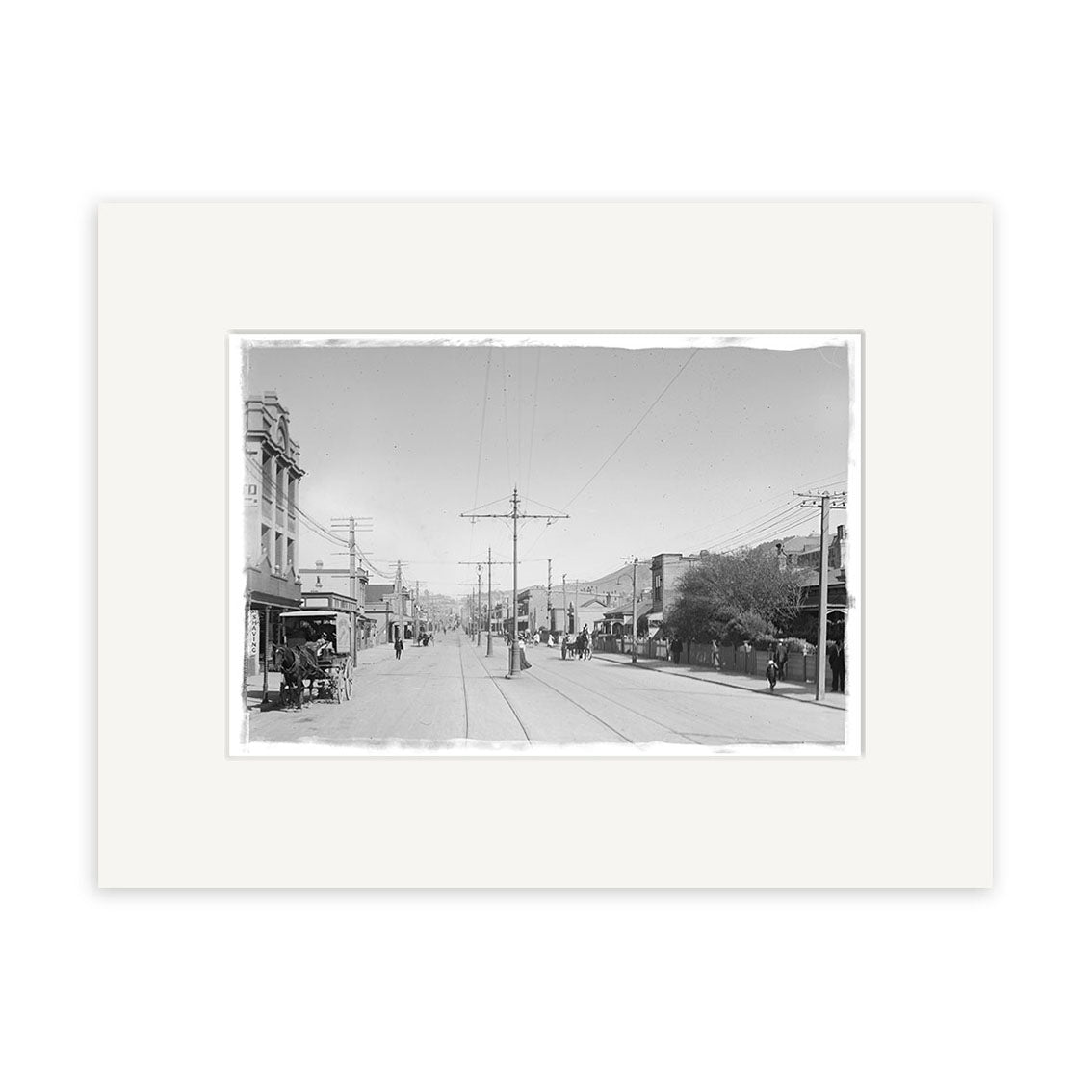 Archival quality black-and-white photographic print: Courtenay Place 1900 No.2 - HI007 shows a New Zealand city street with tram line, utility poles, buildings, and pedestrians, framed by a white mat for preservation.