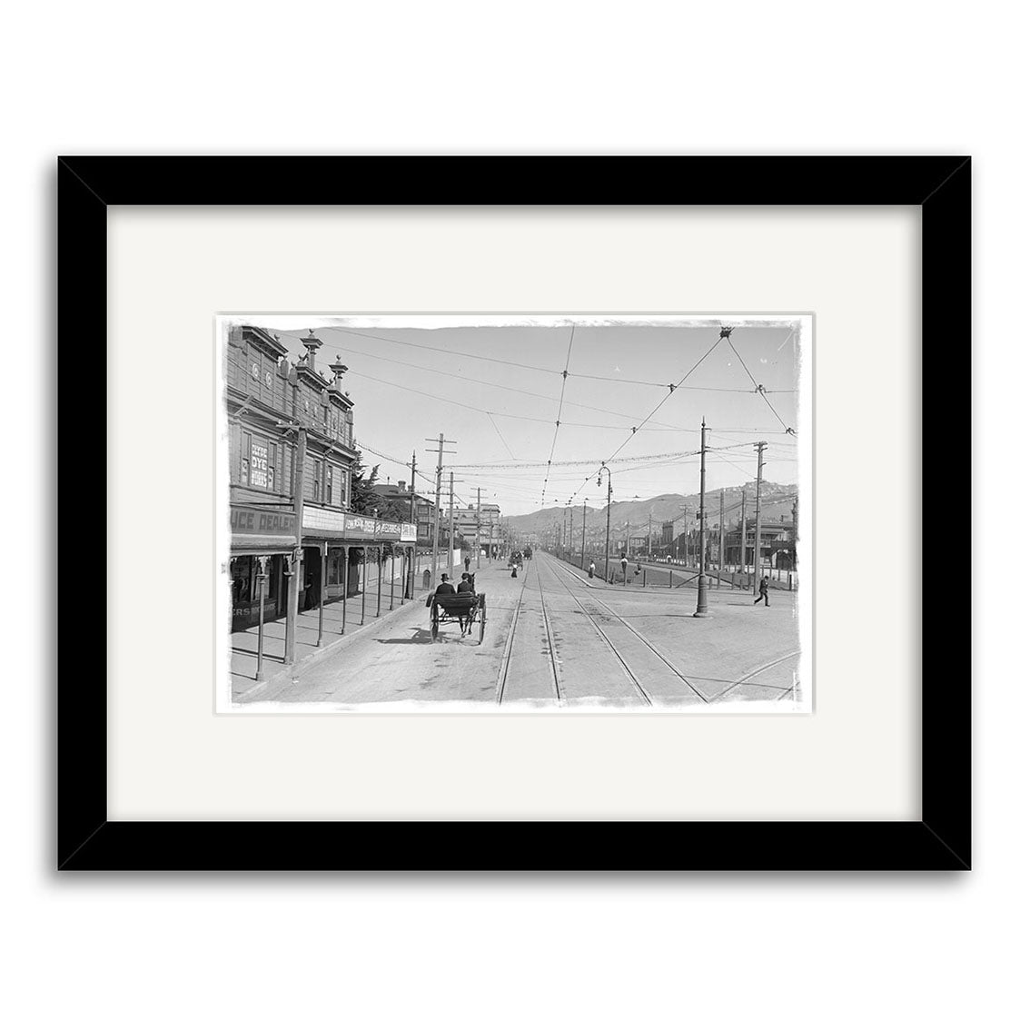 Framed black-and-white photographic print, Kent Terrace 1900 - HI008: Early 20th-century New Zealand street with horse-drawn carriage, trolley tracks, storefronts, and hills. Printed on archival quality matte paper.