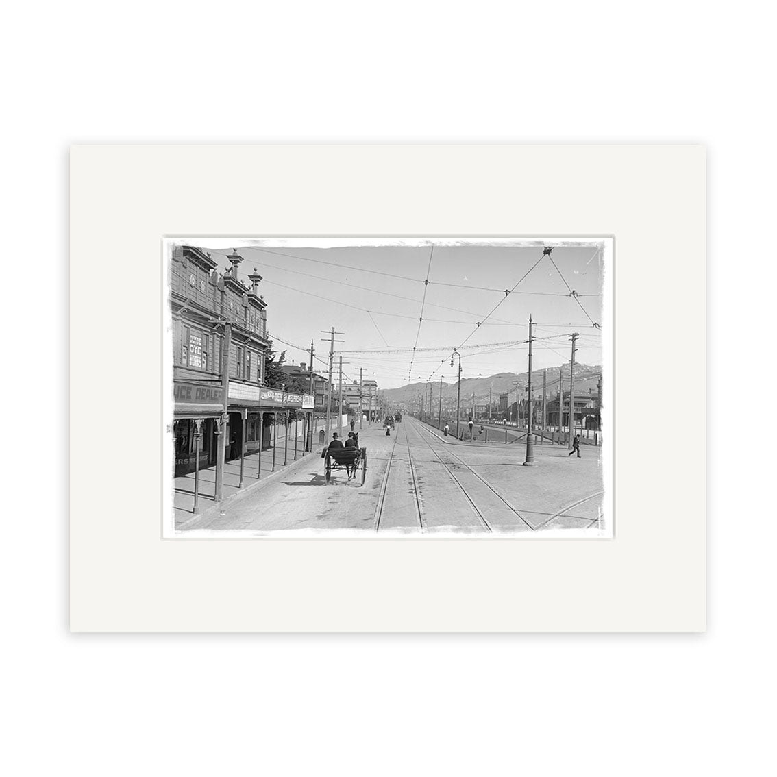 The Kent Terrace 1900 - HI008 is a black and white historical photographic print of a street with trolley tracks, early cars, storefronts, and mountain views. Printed on archival quality matte paper in New Zealand.