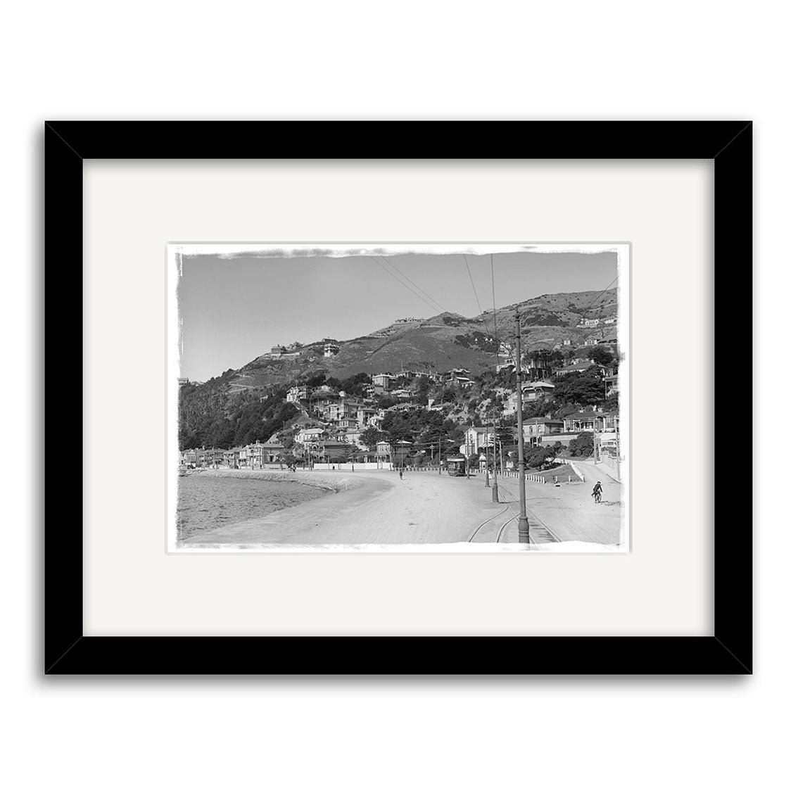 Black-and-white photo Oriental Bay 1900s - HI009 shows a coastal town with hillside houses, sandy beach, and people walking on a paved road; historical print in black frame with white mat, made in New Zealand.