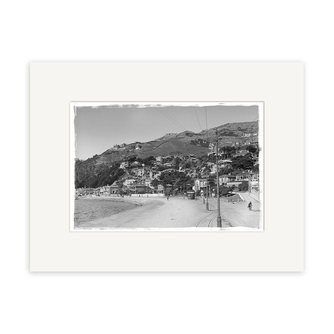 The Oriental Bay 1900s - HI009 is a black and white historical photographic print featuring a coastal town with hillside houses, sandy beach, and people strolling by the shore, framed with a white mat and printed on archival matte paper.